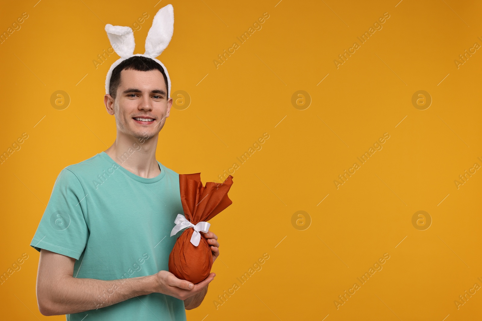Photo of Easter celebration. Handsome young man with bunny ears holding wrapped gift on orange background. Space for text