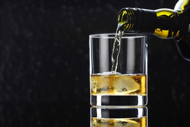 Pouring tasty whiskey from bottle into glass at mirror table against black background, closeup. Space for text