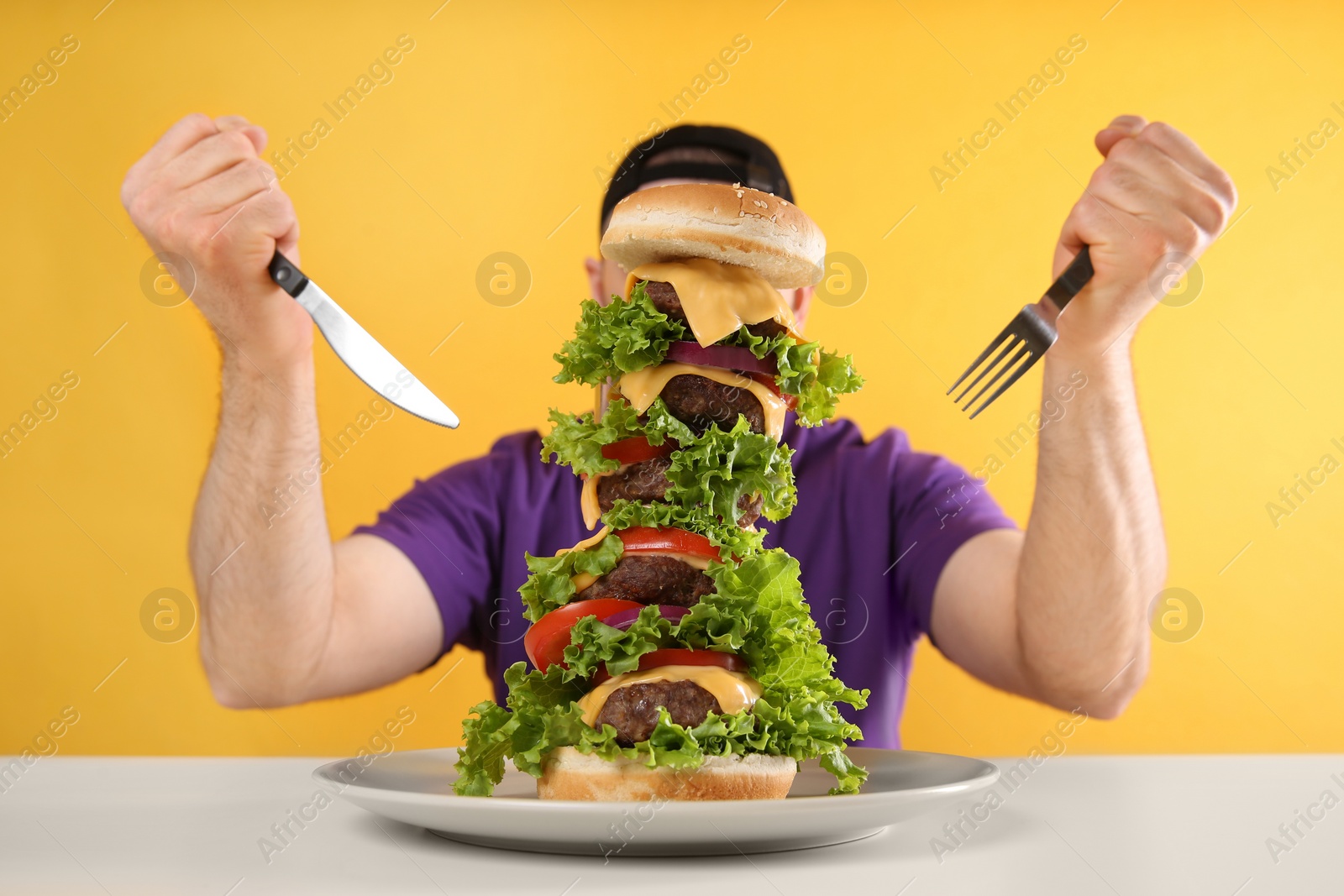 Photo of Hungry man with cutlery and huge burger at white table on yellow background