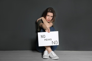 Photo of Young woman holding card with words NO MEANS NO while sitting near grey wall