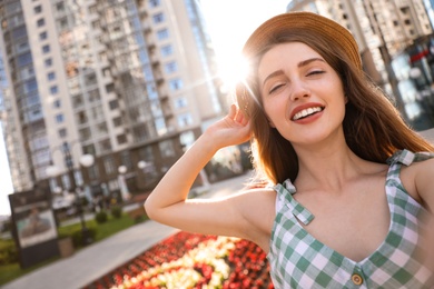 Photo of Beautiful young woman in stylish hat taking selfie outdoors, space for text