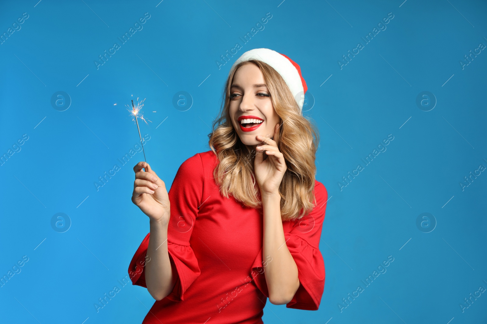 Photo of Happy young woman wearing Santa hat with sparkler on light blue background. Christmas celebration