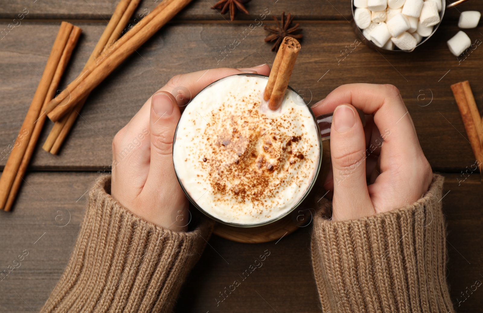Photo of Woman holding cup of hot drink with whipped cream and aromatic cinnamon at wooden table, top view
