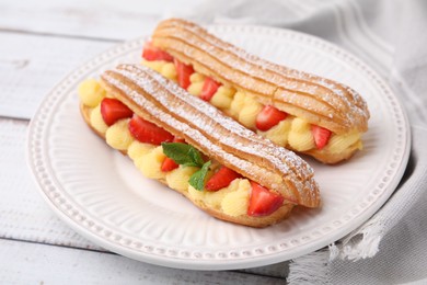 Delicious eclairs filled with cream, strawberries and mint on white wooden table, closeup