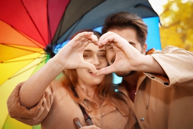 Photo of Happy couple with colorful umbrella making heart with hands in park