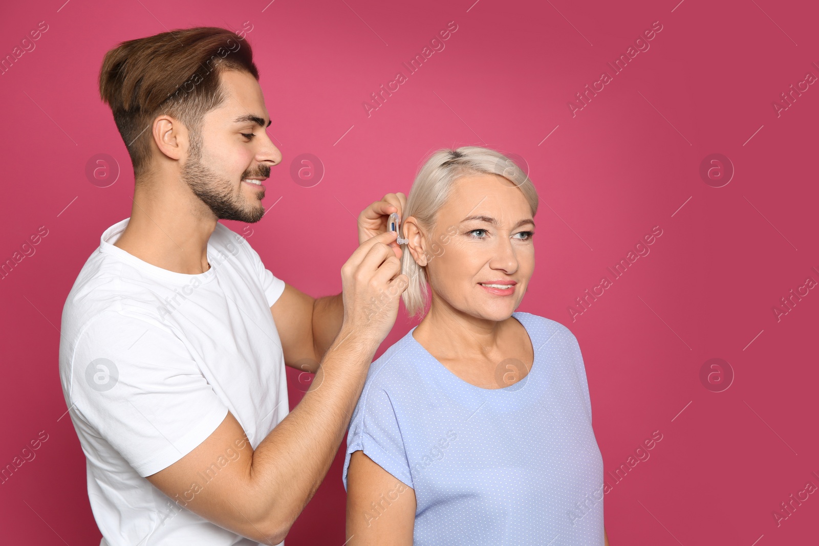 Photo of Young man putting hearing aid in mother's ear on color background