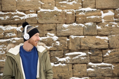 Young man listening to music with headphones against stone wall. Space for text