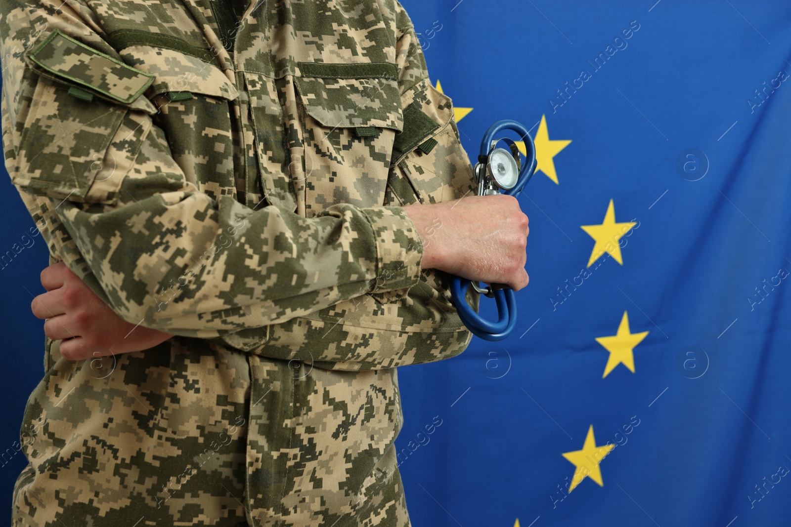 Photo of Man in military uniform with crossed arms and stethoscope against flag of European Union, closeup. Health care concept