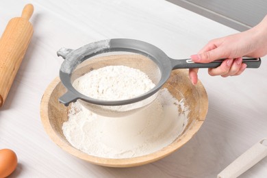 Photo of Woman sieving flour into bowl at white wooden table in kitchen, closeup