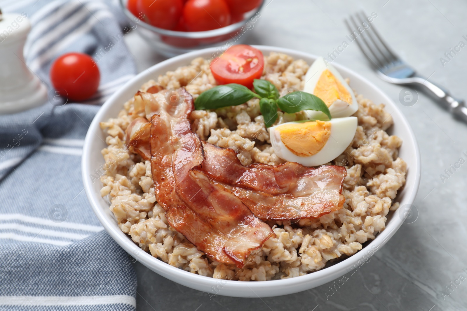 Photo of Tasty boiled oatmeal with egg, bacon and tomatoes served on light grey table, closeup