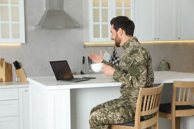 Photo of Soldier with cup of drink using video chat on laptop at white marble table in kitchen. Military service