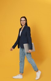 Young woman with modern laptop walking on yellow background