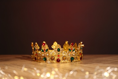 Photo of Beautiful golden crown with gems and fairy lights on wooden table. Fantasy item