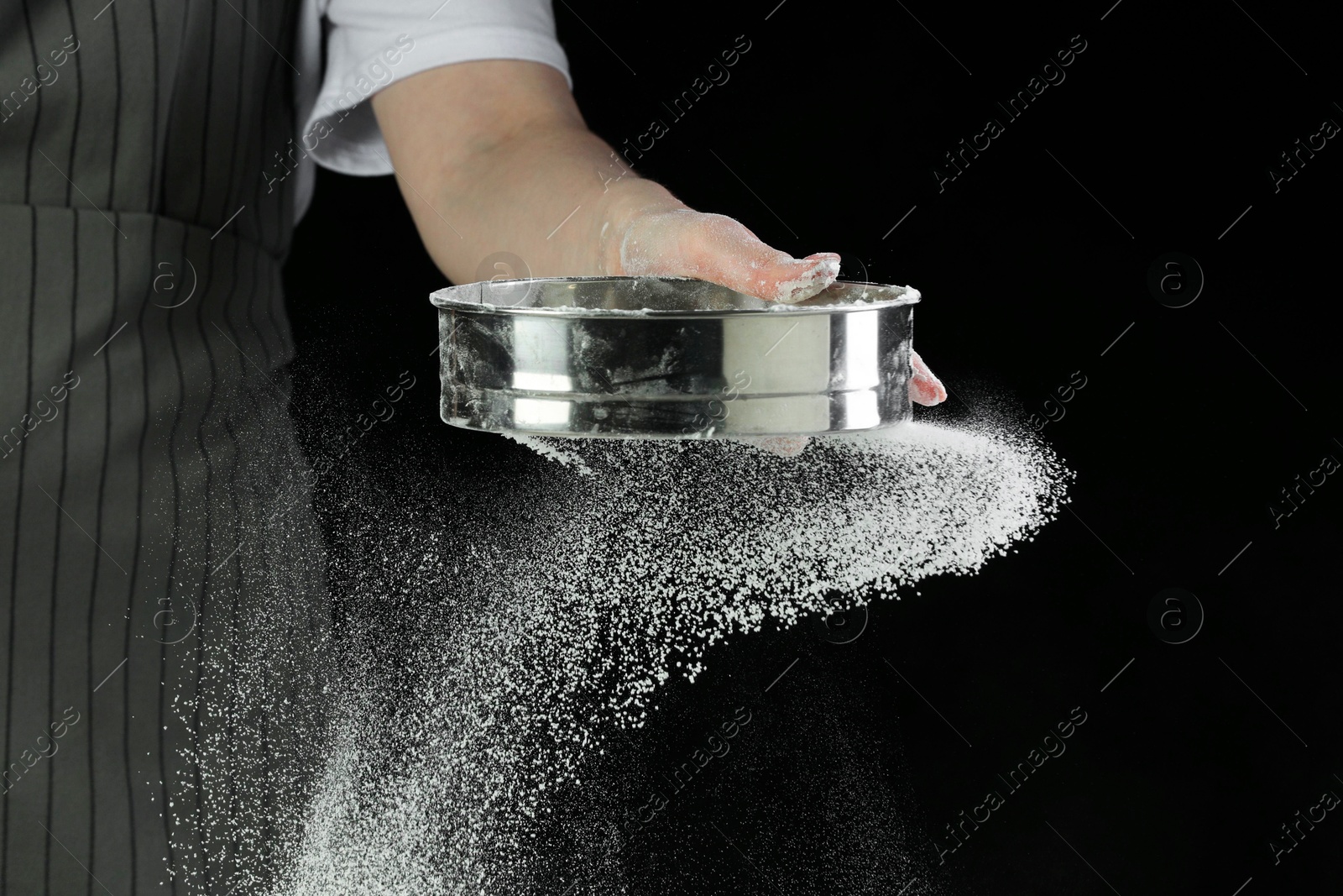 Photo of Woman sieving flour at table against black background, closeup