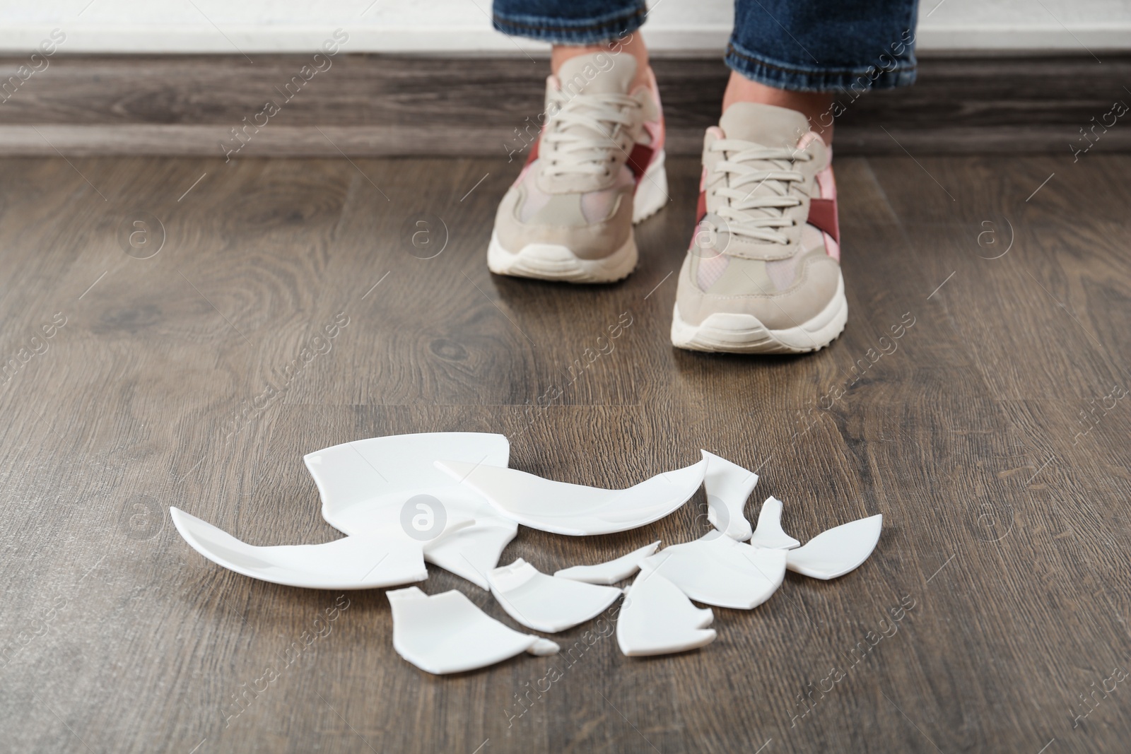 Photo of Woman in sneakers standing near broken plate on floor indoors, closeup