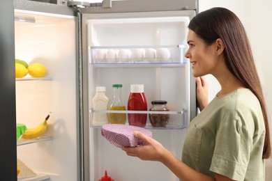 Photo of Happy woman holding bowl covered with beeswax food wrap near refrigerator in kitchen