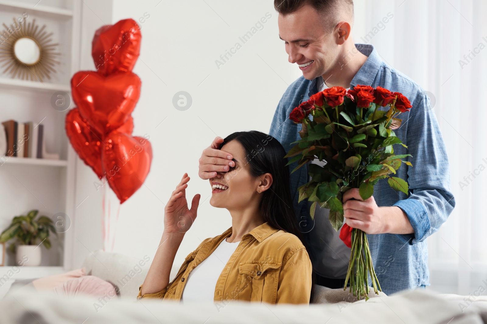 Photo of Boyfriend presenting beautiful bouquet of roses to his girlfriend in room decorated with heart shaped balloons. Valentine's day celebration