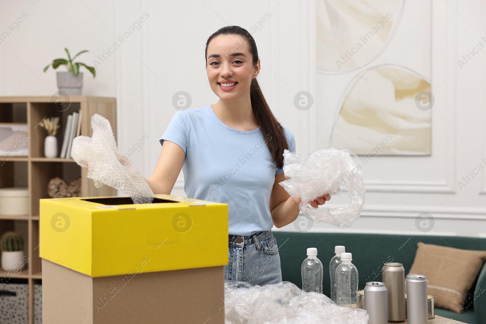 Photo of Garbage sorting. Smiling woman throwing plastic package into cardboard box in room