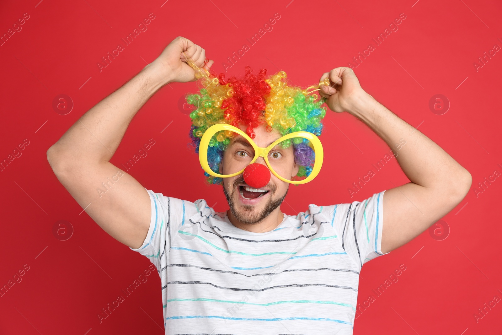 Photo of Funny man with large glasses, rainbow wig and clown nose on red background. April fool's day