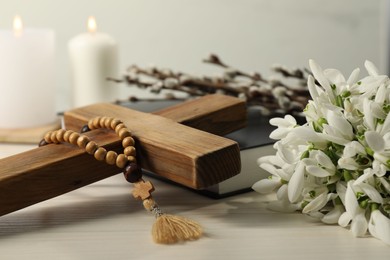 Wooden cross, rosary beads, Bible, church candles and snowdrops on white table, closeup