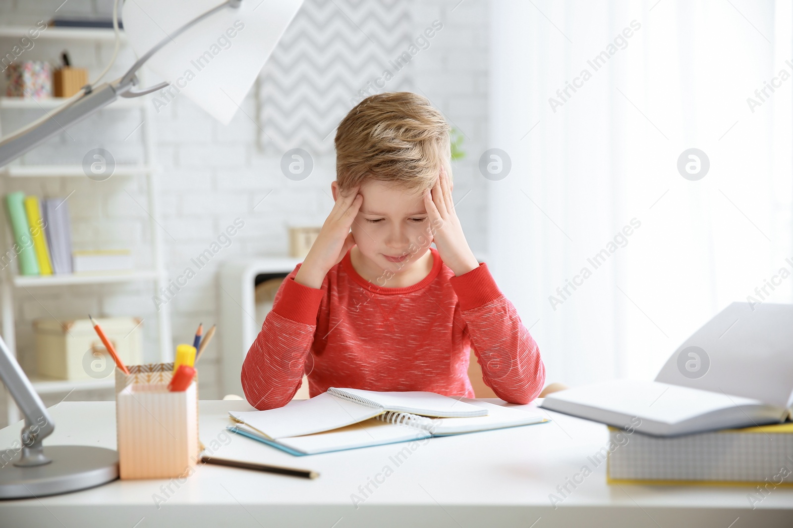 Photo of Little boy suffering from headache while doing homework at table indoors