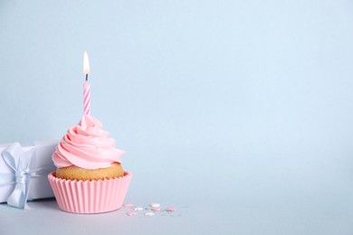 Delicious birthday cupcake with burning candle, sprinkles and gift box on light blue background, space for text