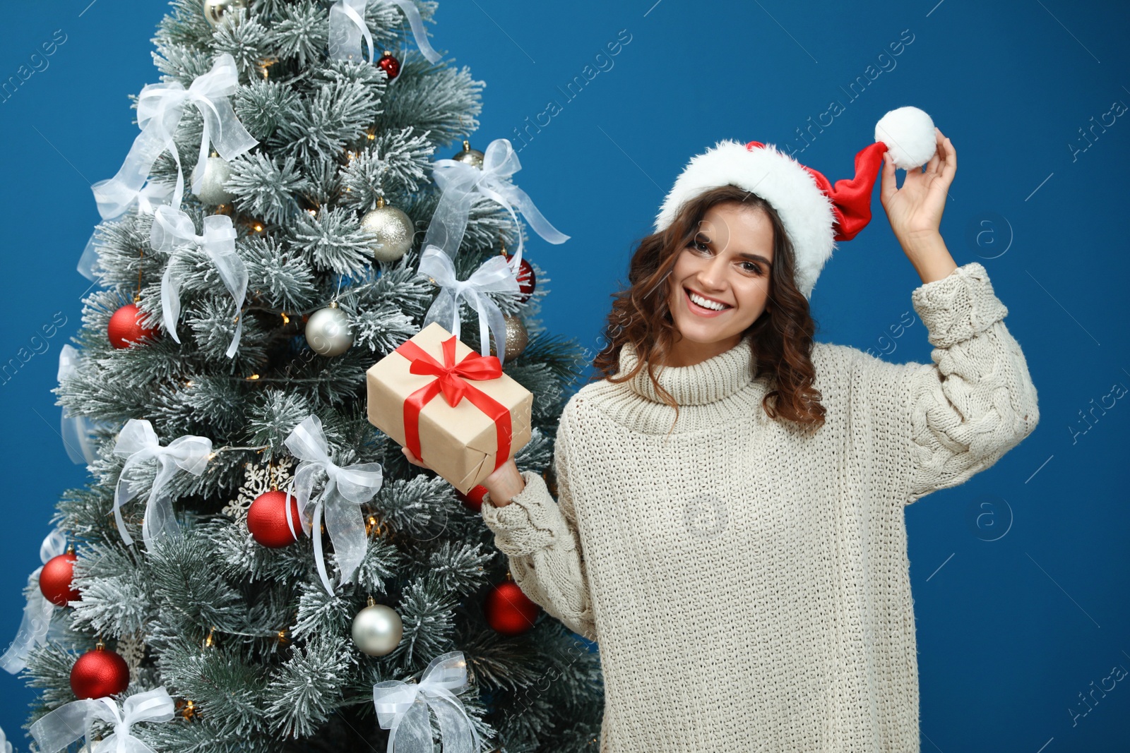 Image of Happy young woman in Santa hat with gift near Christmas tree on blue background