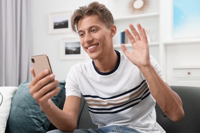 Happy young man having video chat via smartphone on sofa indoors