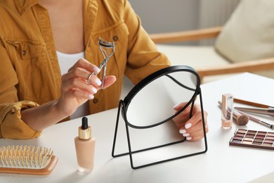 Woman with eyelash curler, makeup products and mirror at table indoors, closeup