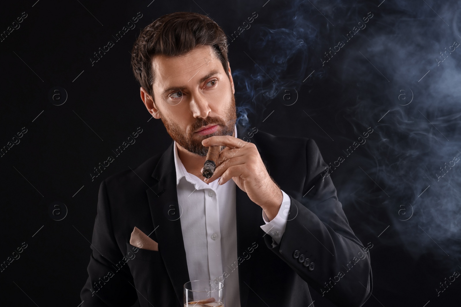 Photo of Handsome man in elegant suit with glass of whiskey smoking cigar on black background