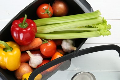 Photo of Black pot with fresh vegetables and glass lid on white wooden table, top view