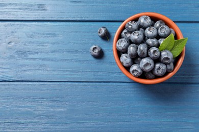 Photo of Tasty fresh blueberries with green leaves in bowl on blue wooden table, flat lay. Space for text
