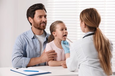 Doctor examining girl`s oral cavity with tongue depressor near her father indoors