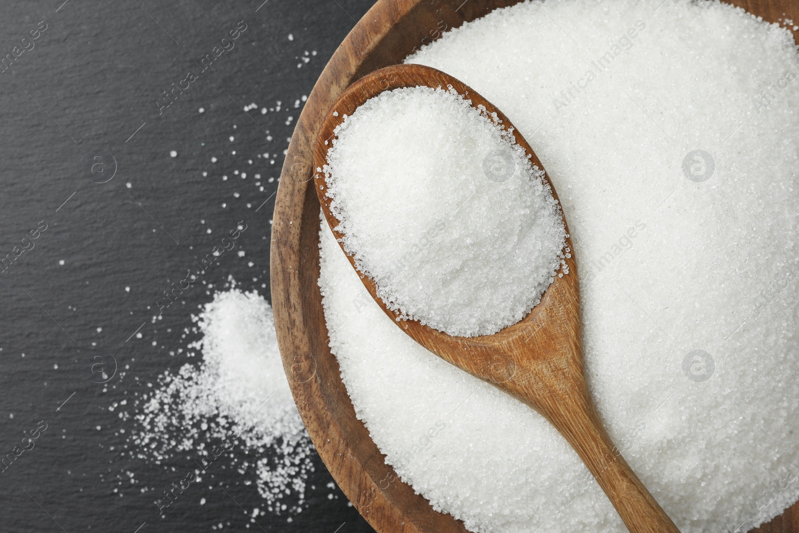 Photo of Granulated sugar in bowl and spoon on black table, top view. Space for text