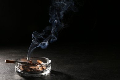 Smoldering cigarette in glass ashtray on grey table against black background. Space for text