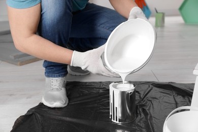 Photo of Man pouring white paint from bucket into can indoors, closeup