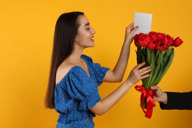 Photo of Happy woman receiving red tulip bouquet with card from man on yellow background. 8th of March celebration