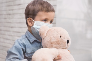 Sad little boy in protective mask looking out of window indoors, view from outside. Staying at home during coronavirus pandemic