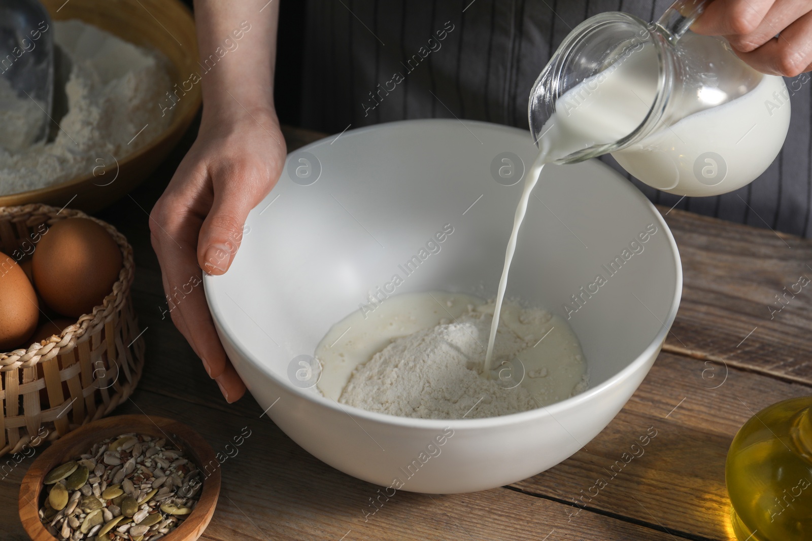 Photo of Making bread. Woman pouring milk into bowl at wooden table, closeup