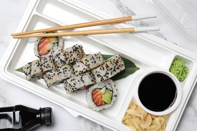 Food delivery. Delicious sushi rolls with soy sauce, ginger, wasabi and chopsticks in plastic container on white marble table, flat lay