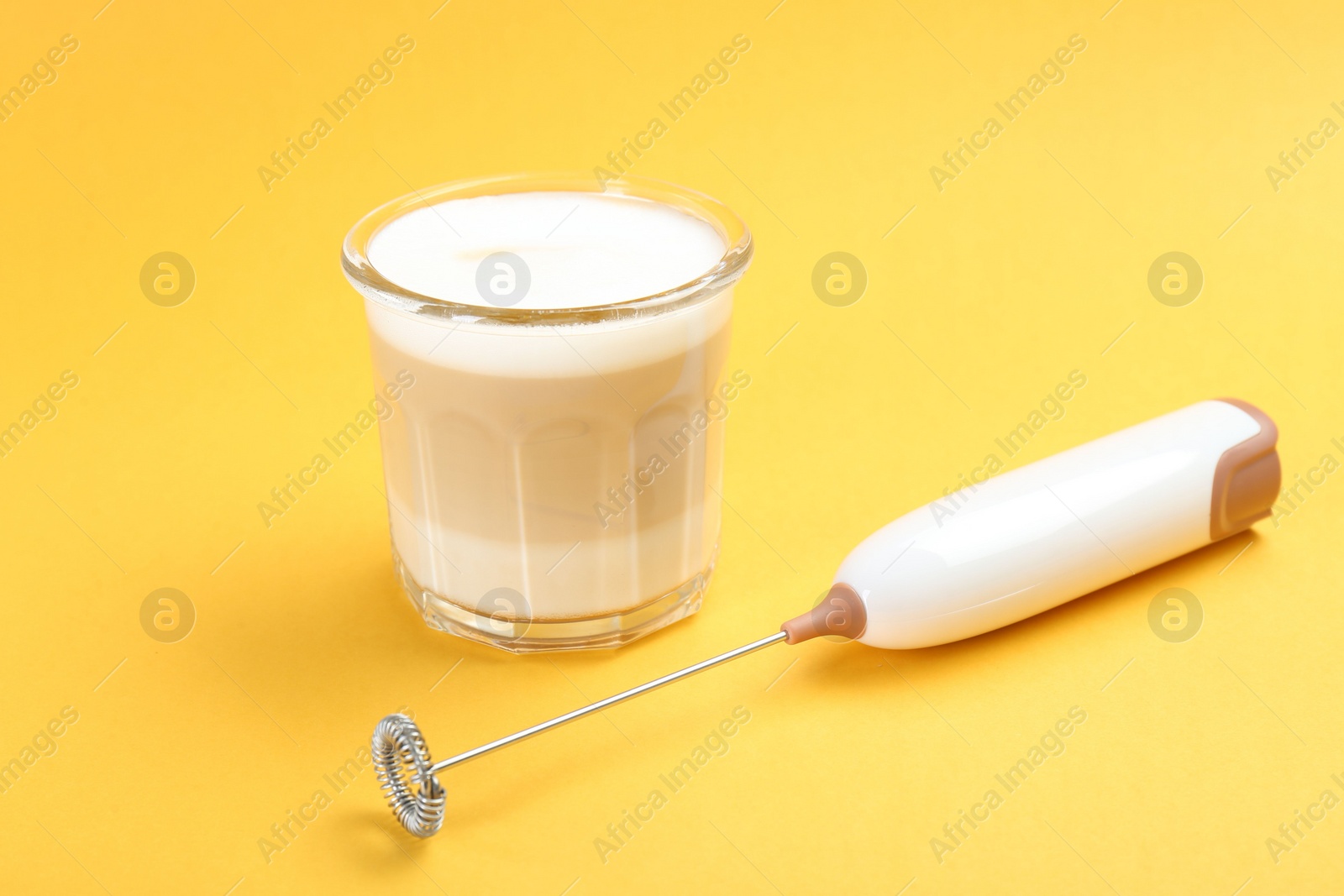 Photo of Mini mixer (milk frother) and tasty cappuccino in glass on yellow background