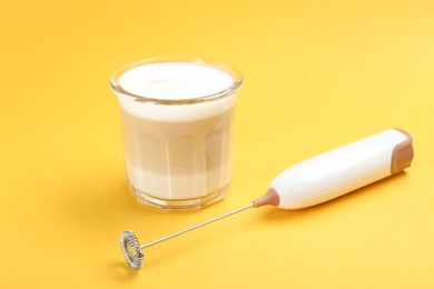 Mini mixer (milk frother) and tasty cappuccino in glass on yellow background