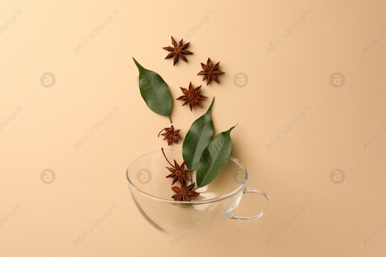 Photo of Anise stars and green leaves falling into glass cup on beige background, flat lay