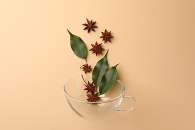 Anise stars and green leaves falling into glass cup on beige background, flat lay