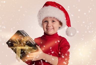 Image of Cute child in Santa hat with Christmas gift on light background