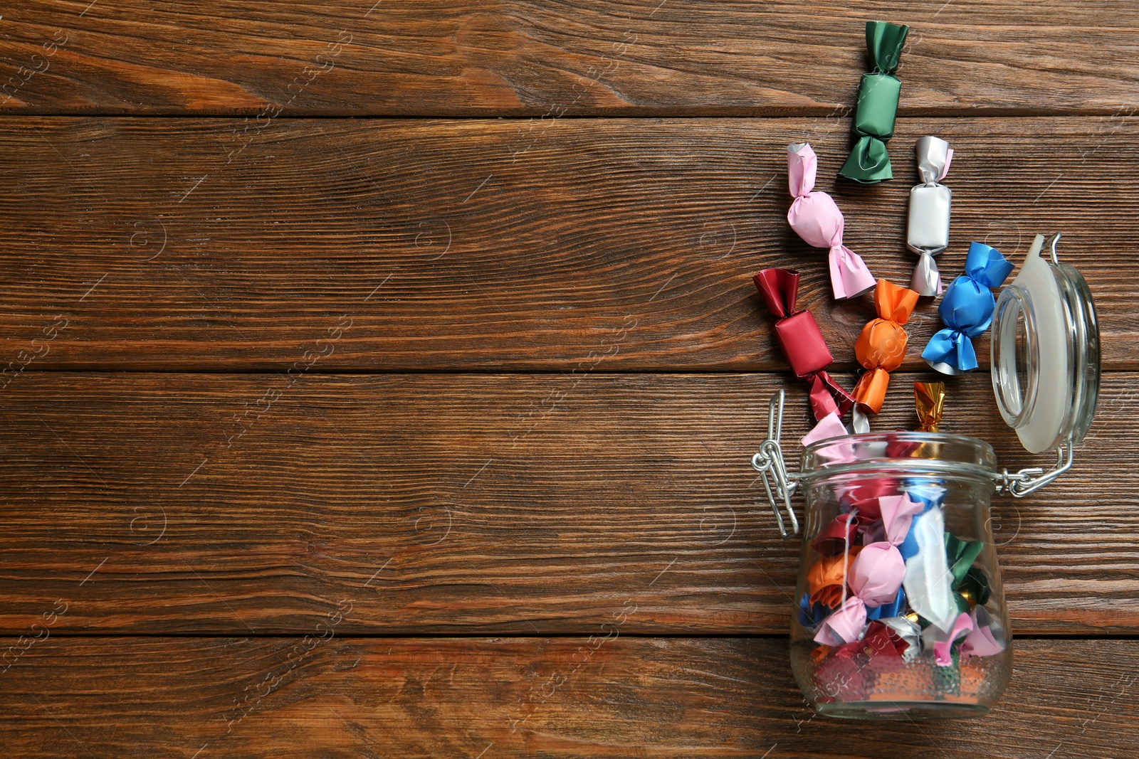 Photo of Overturned jar with candies in colorful wrappers on wooden table, flat lay. Space for text