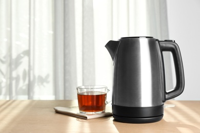 Photo of Modern electric kettle and cup of tea on wooden table indoors. Space for text