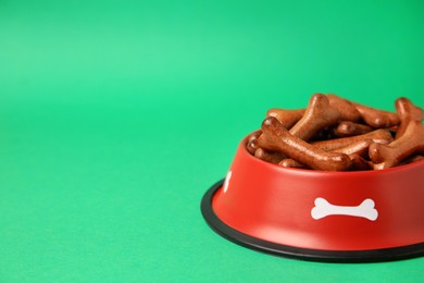 Red bowl with bone shaped dog cookies on green background. Space for text