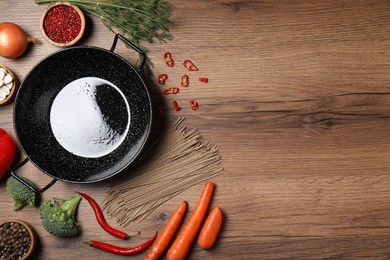 Empty iron wok surrounded by raw ingredients on wooden table, flat lay. Space for text