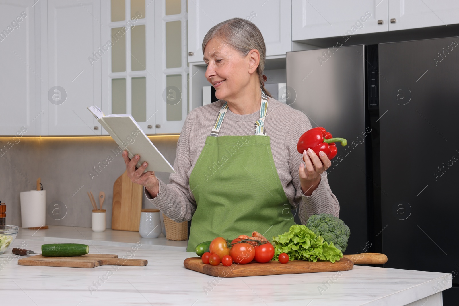 Photo of Happy woman with recipe book and bell pepper in kitchen